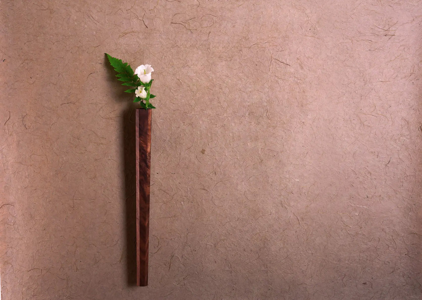 A solid walnut wall vase with a small white flower against a brown wall.