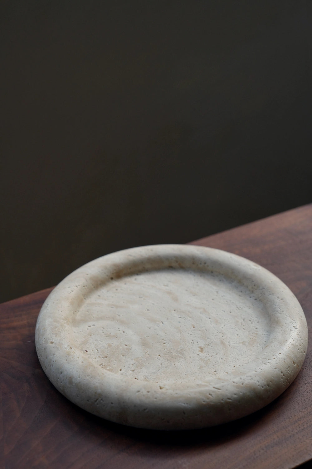 The round travertine catchall tray resting on the corner of a dark wood surface, showcasing its minimalist and sculptural form.