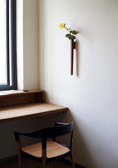 A long, octagonal wood wall vase with a yellow flower, near a desk.