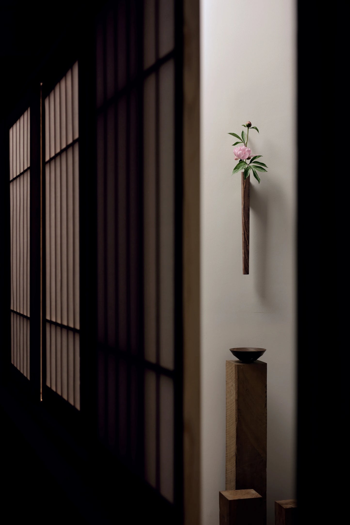 A solid walnut Ikebana-style wall vase with a pink peony in a dark room.