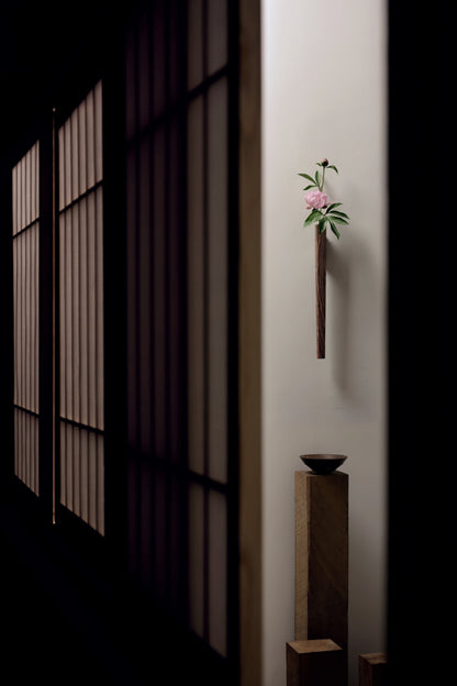 A solid walnut Ikebana-style wall vase with a pink peony in a dark room.