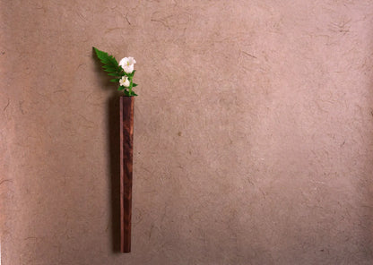 A solid walnut wall vase with a small white flower against a brown wall.