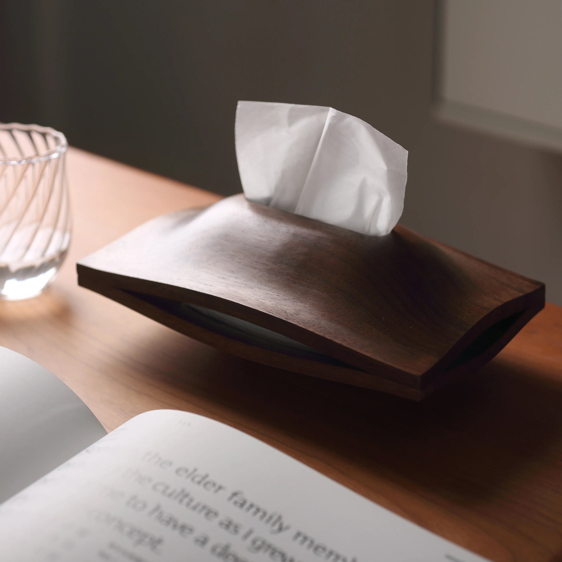 A walnut tissue holder on a desk next to an open book.