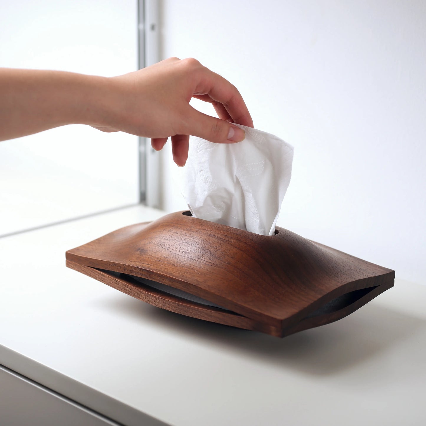 Hand pulling a fresh tissue from the walnut sculptural holder.
