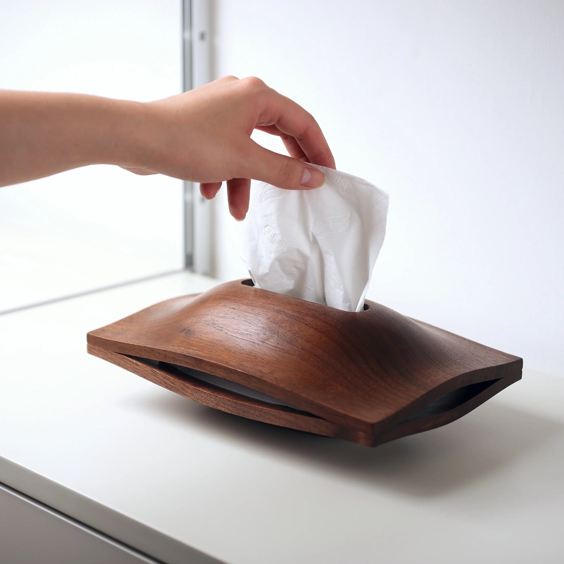 Hand pulling a fresh tissue from the walnut sculptural holder.