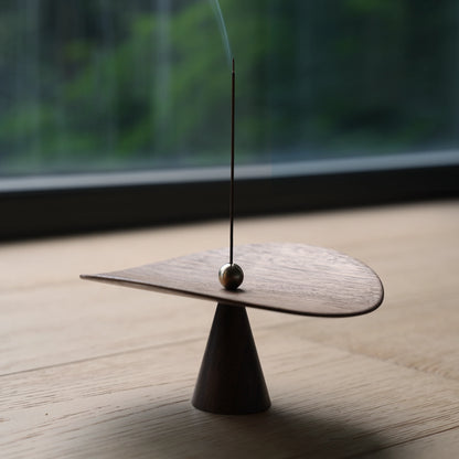 Modern walnut incense holder on a wood table with a window behind.
