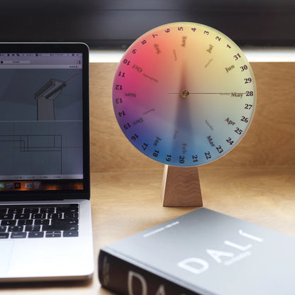 Chromatic perpetual calendar on a desk next to a laptop and book.