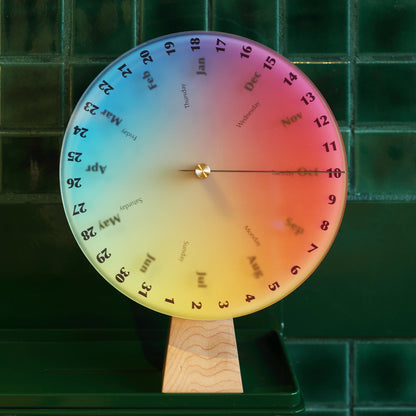 Rainbow perpetual calendar sitting on a shelf against green tile.