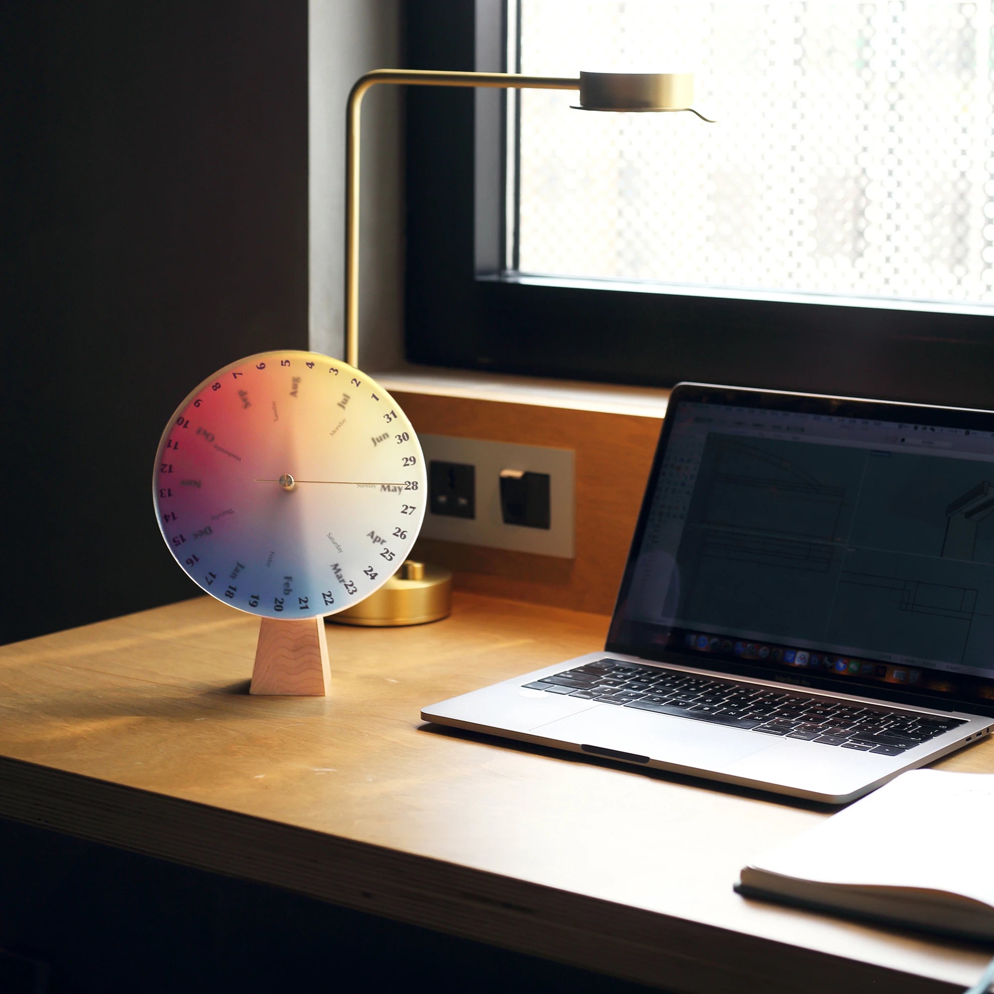 A perpetual desk calendar next to a laptop and a modern gold lamp.