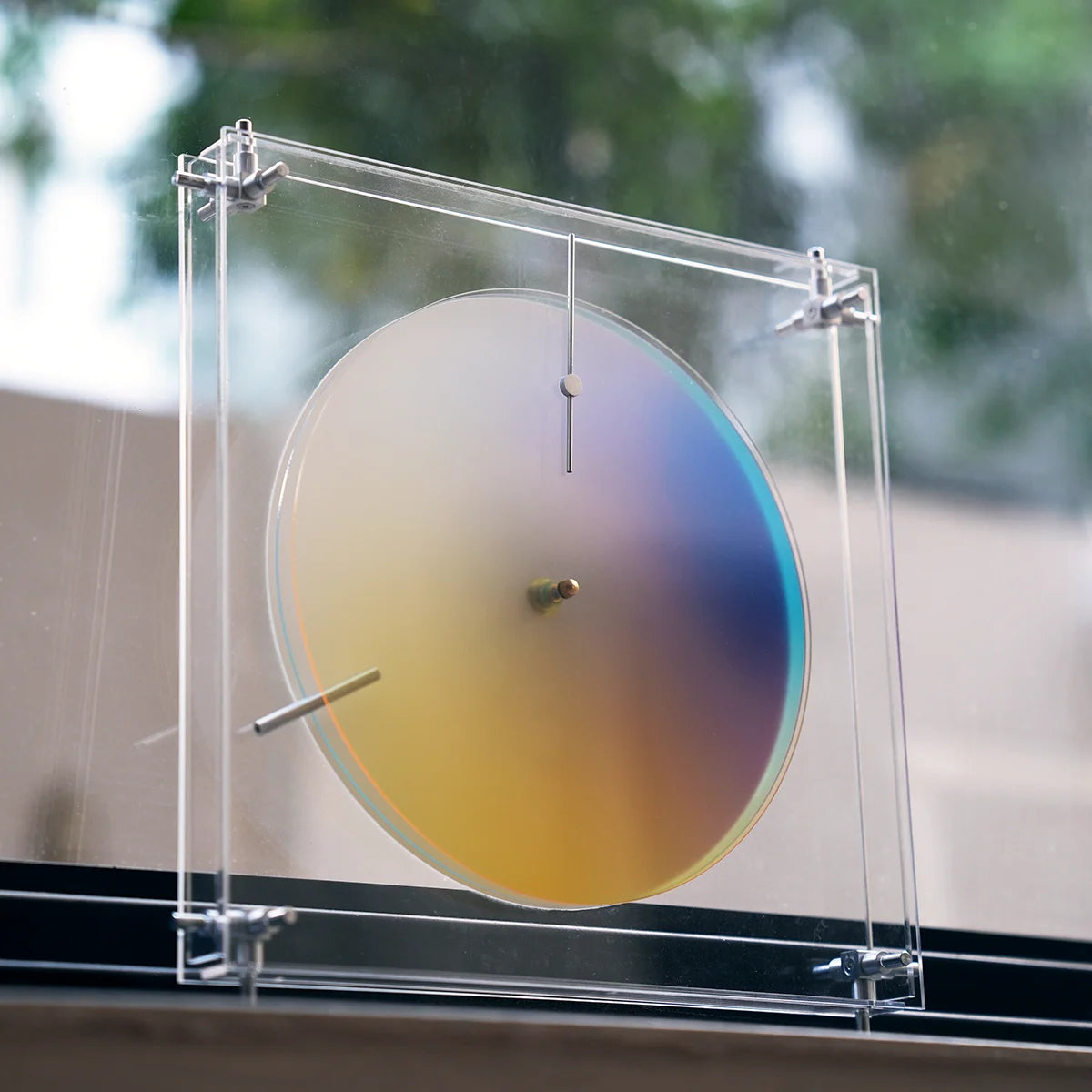 Chromatic gradient wall clock in a clear acrylic frame by a window.