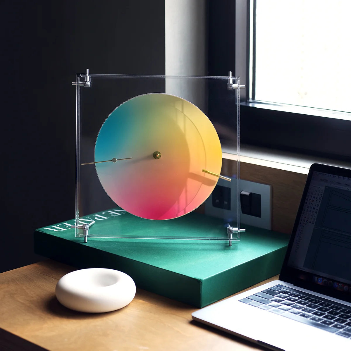 A modern color wheel clock standing on a book next to a laptop.