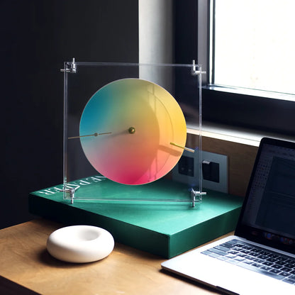 A modern color wheel clock standing on a book next to a laptop.