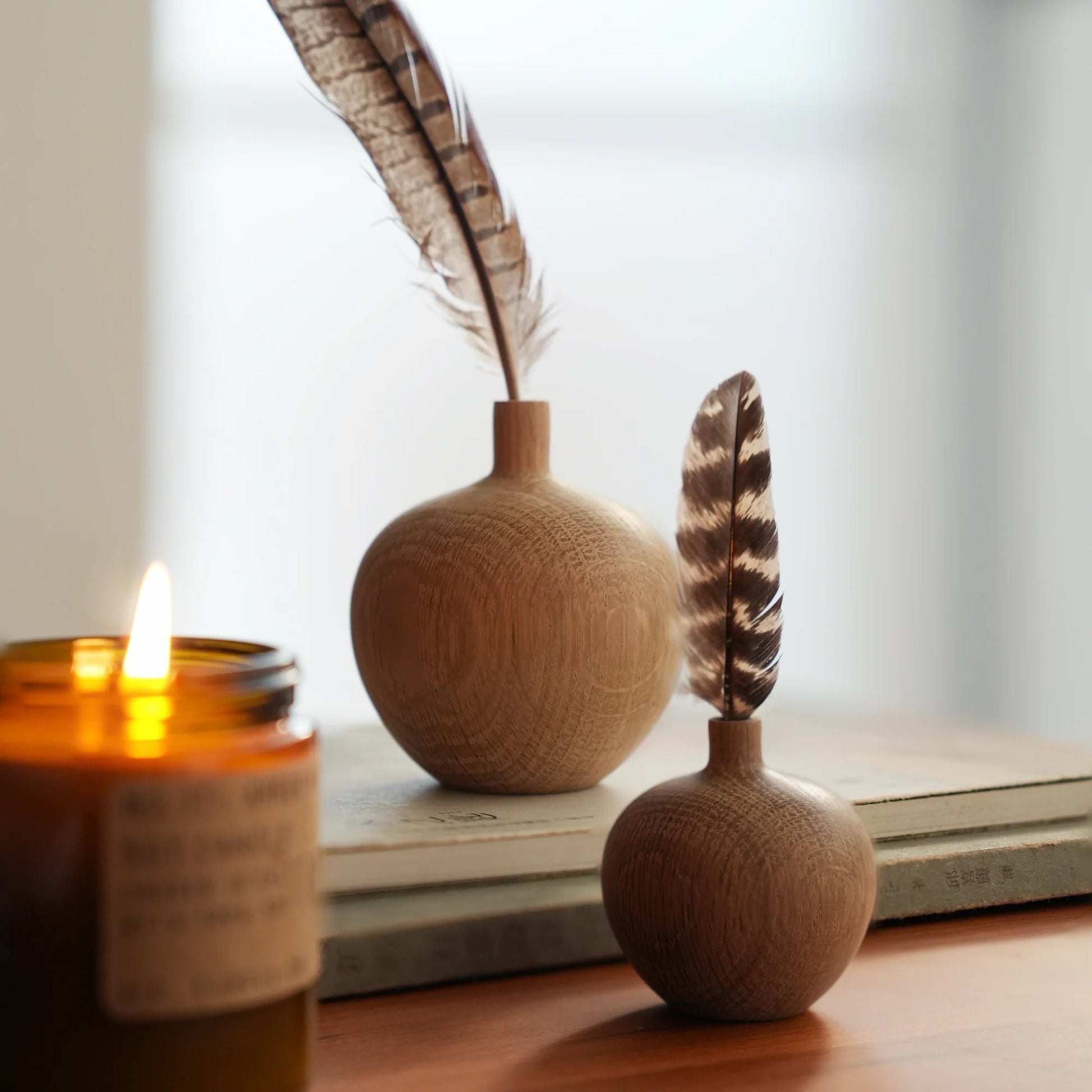 Two oak bud vases with feathers styled on a desk with a candle.