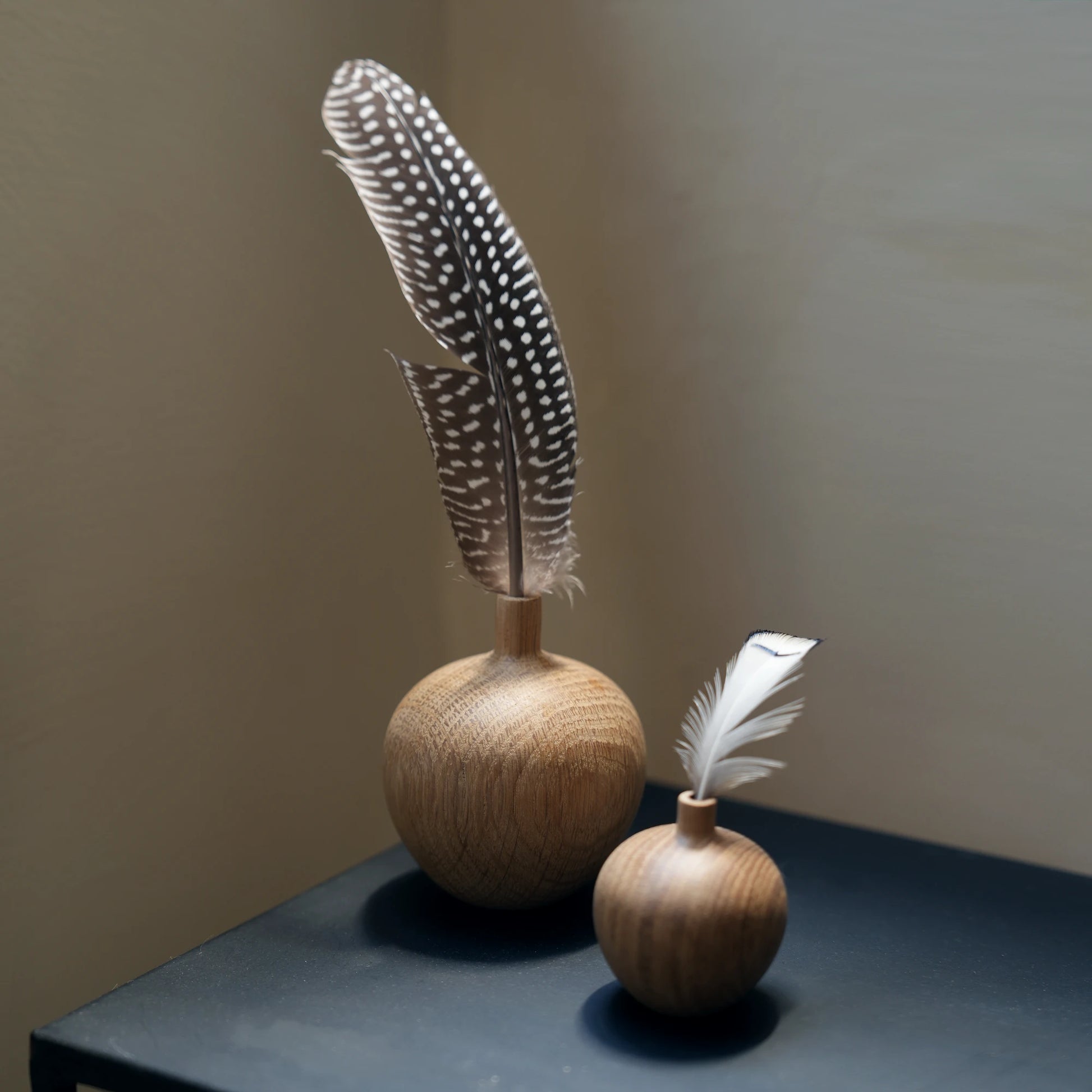 Two oak vases with spotted guinea fowl feathers on a black table.