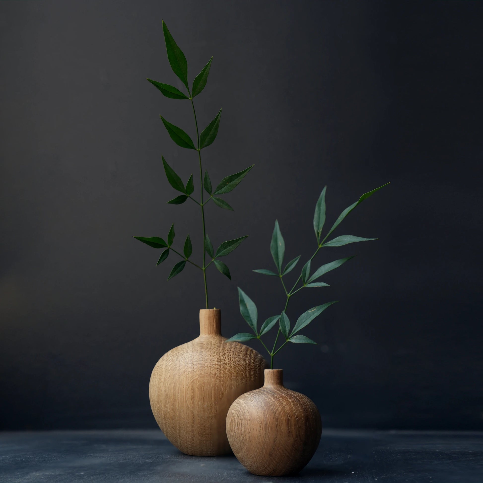 A pair of oak wood bud vases with green stems against a dark background.