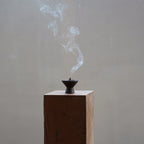 Plume of smoke rising from a cone incense burner on a wood block.