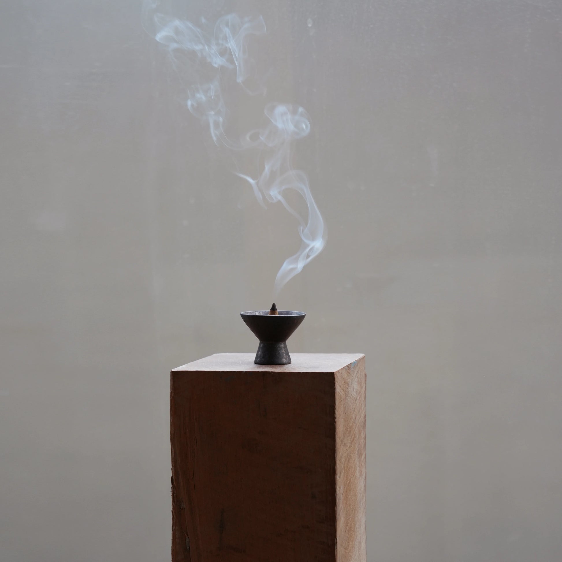 Plume of smoke rising from a cone incense burner on a wood block.