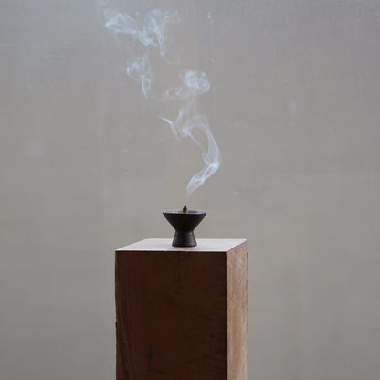 Plume of smoke rising from a cone incense burner on a wood block.