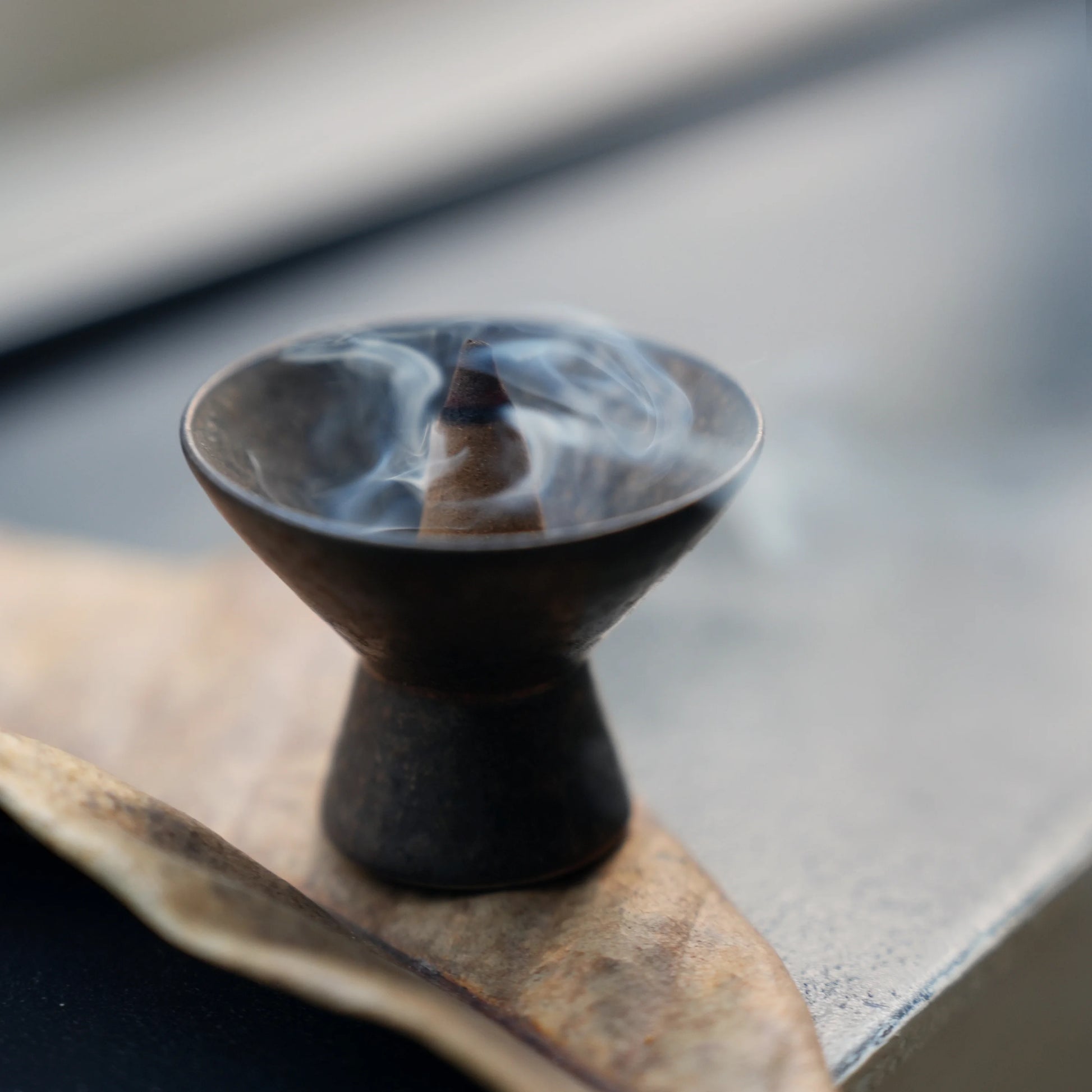 Smoke from an incense cone in a ceramic holder resting on a leaf.