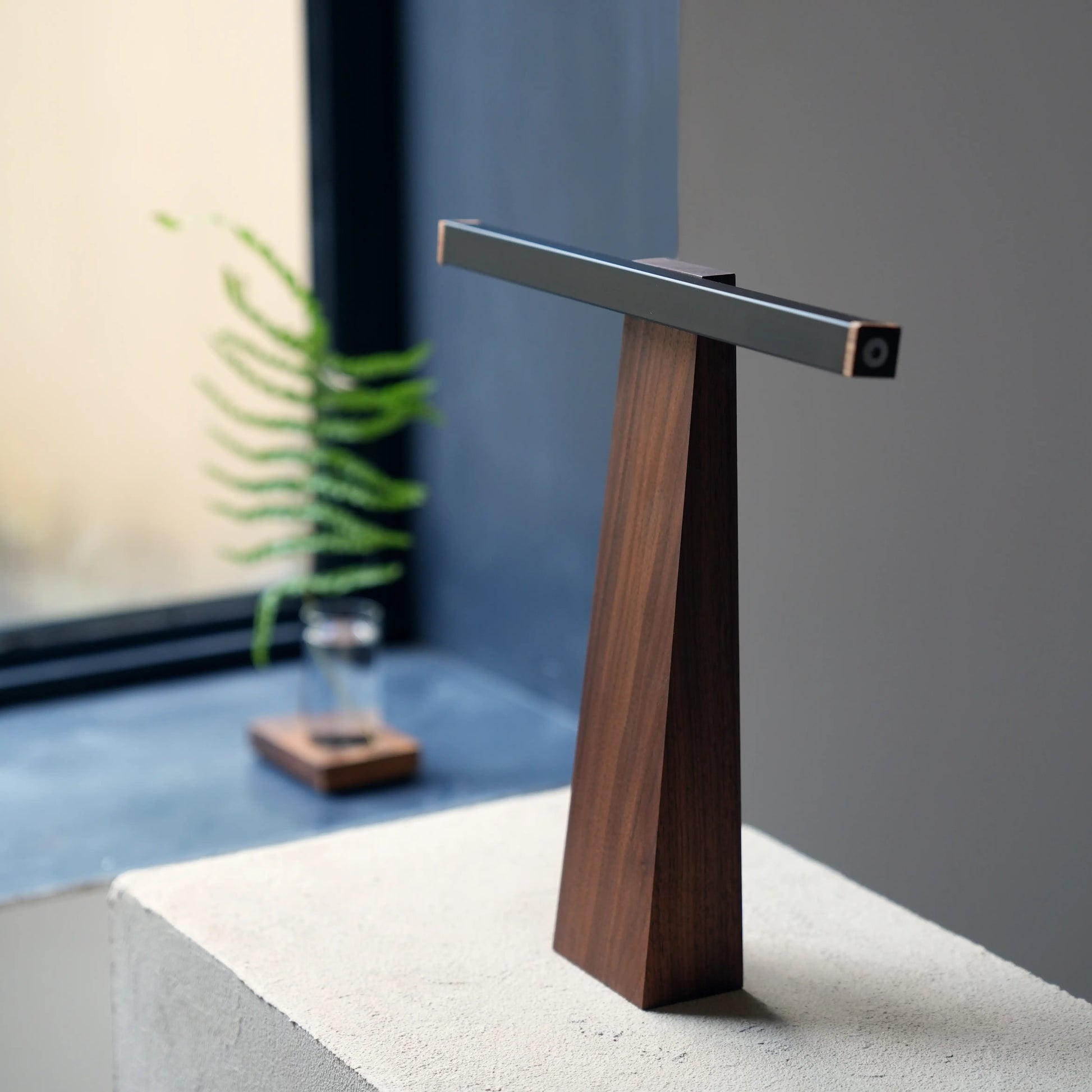 Sculptural T-shaped LED desk lamp in walnut and black aluminum on a white block.