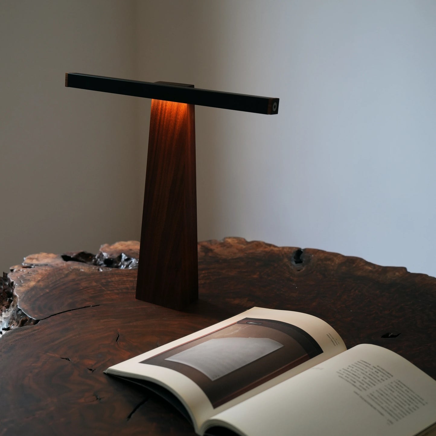 The sculptural walnut and aluminum lamp rests on a dark, live-edge wood table, its integrated LED casting a warm, inviting glow. The light illuminates an open art book in the foreground, highlighting the rich, textured grain of the wood for a cozy reading nook.