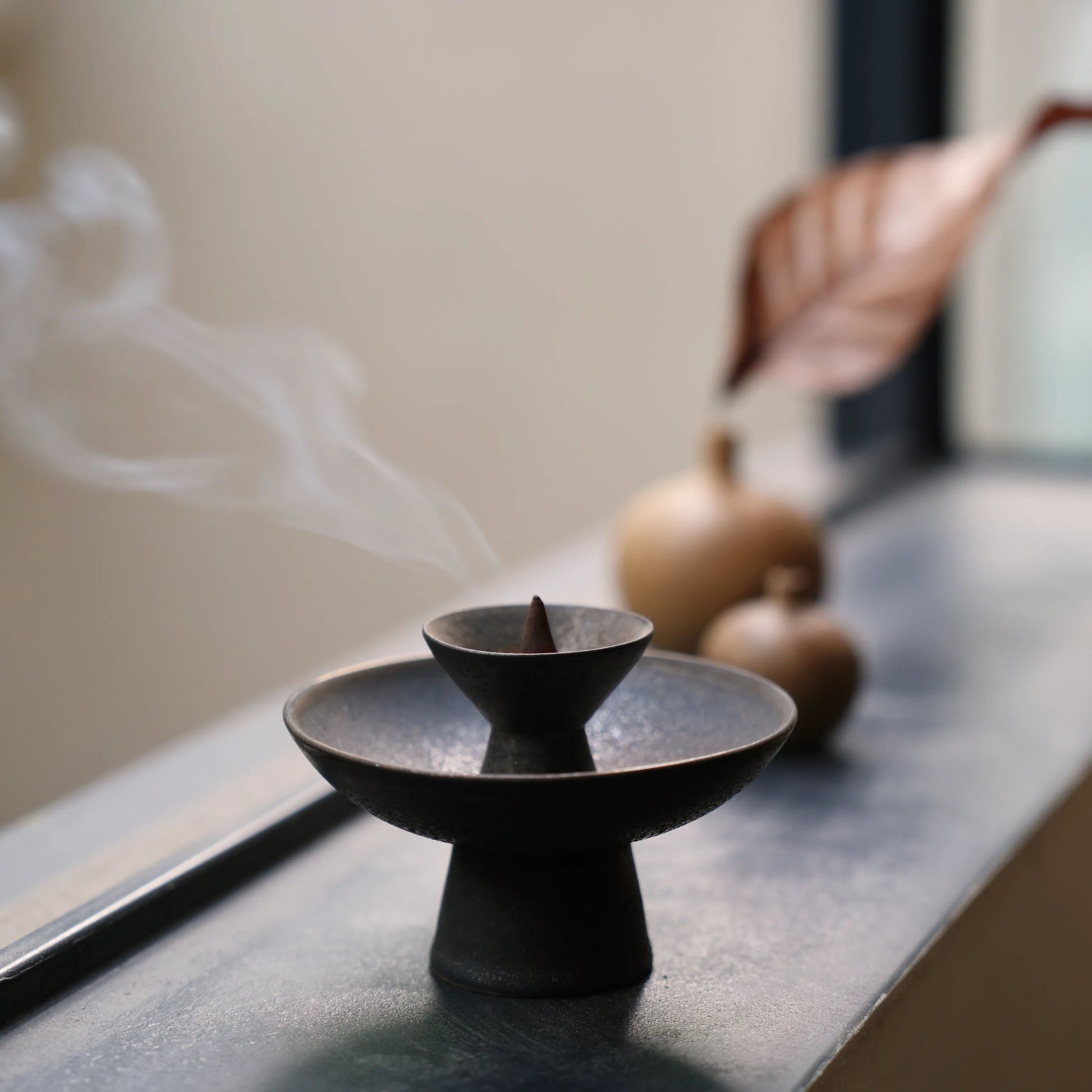 Smoke rising from incense cone in a black ceramic burner on windowsill.