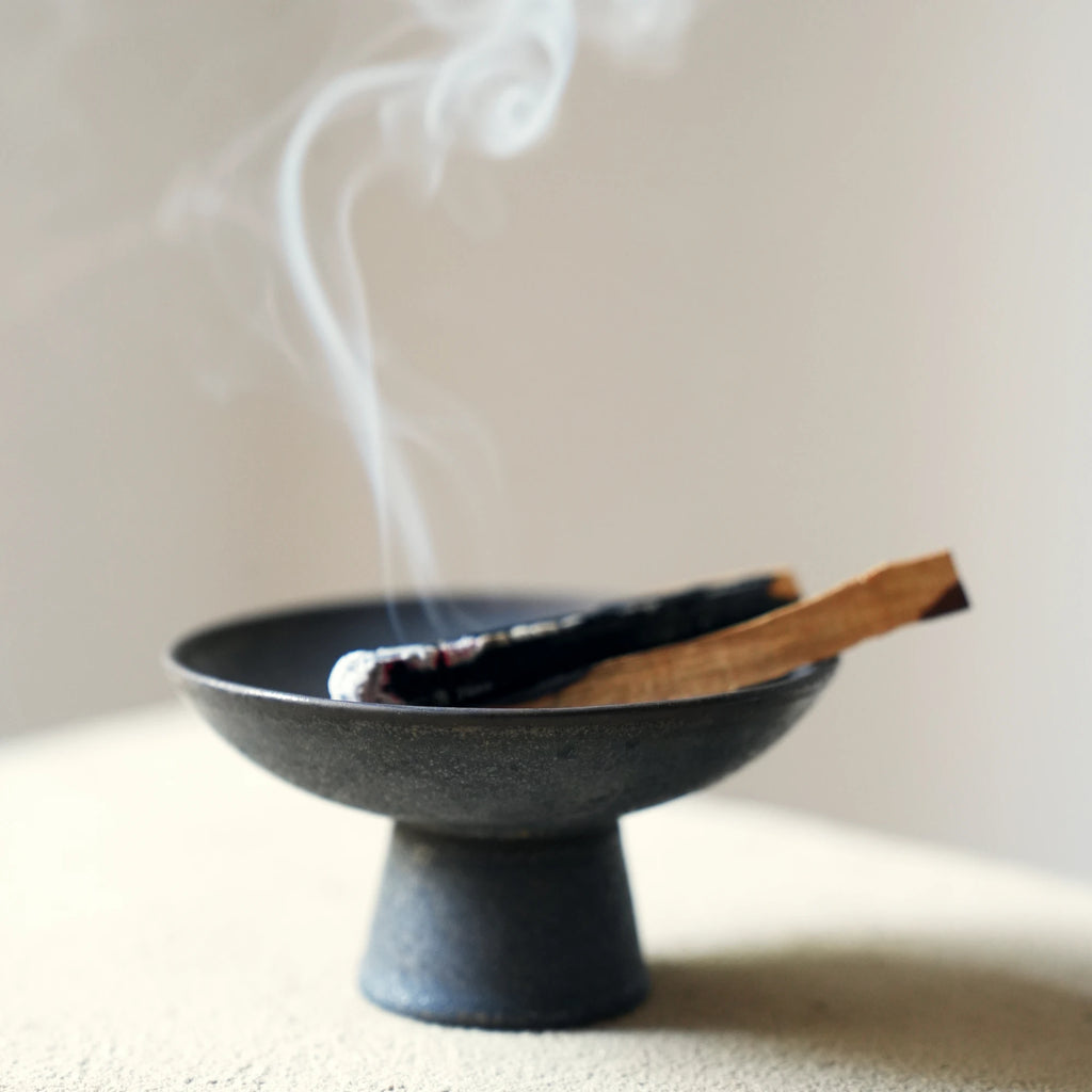 Smoke curls from a burning Palo Santo stick in a ceramic dish.