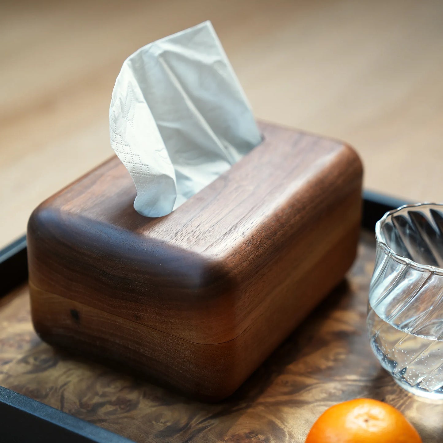 A dark wood tissue box on a tray with a glass and a tangerine.