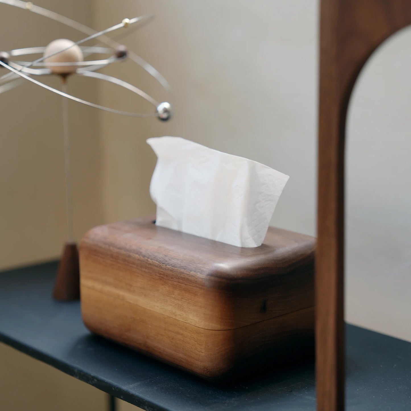 A walnut tissue box next to a kinetic sculpture on a black shelf.