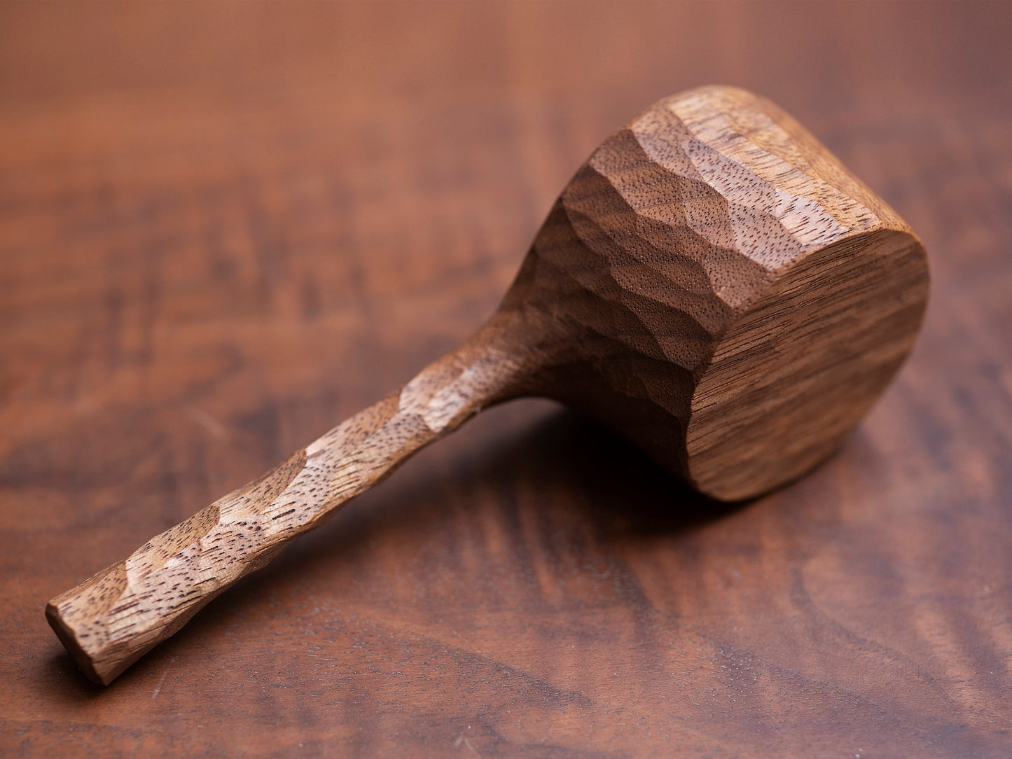 A hand-carved, faceted walnut coffee scoop resting on a dark wood table.