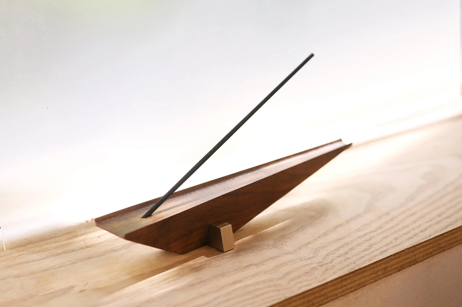 A modern walnut and brass incense holder sitting on a light wood windowsill.