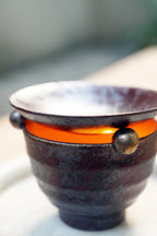 Close-up: dark ceramic oil burner, bronze knobs, orange candle glow
