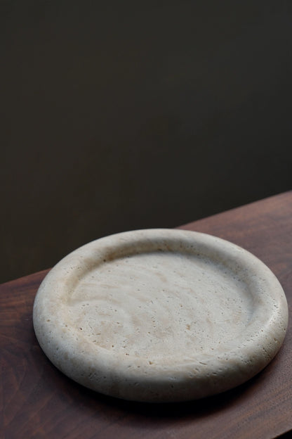 The round travertine catchall tray resting on the corner of a dark wood surface, showcasing its minimalist and sculptural form.