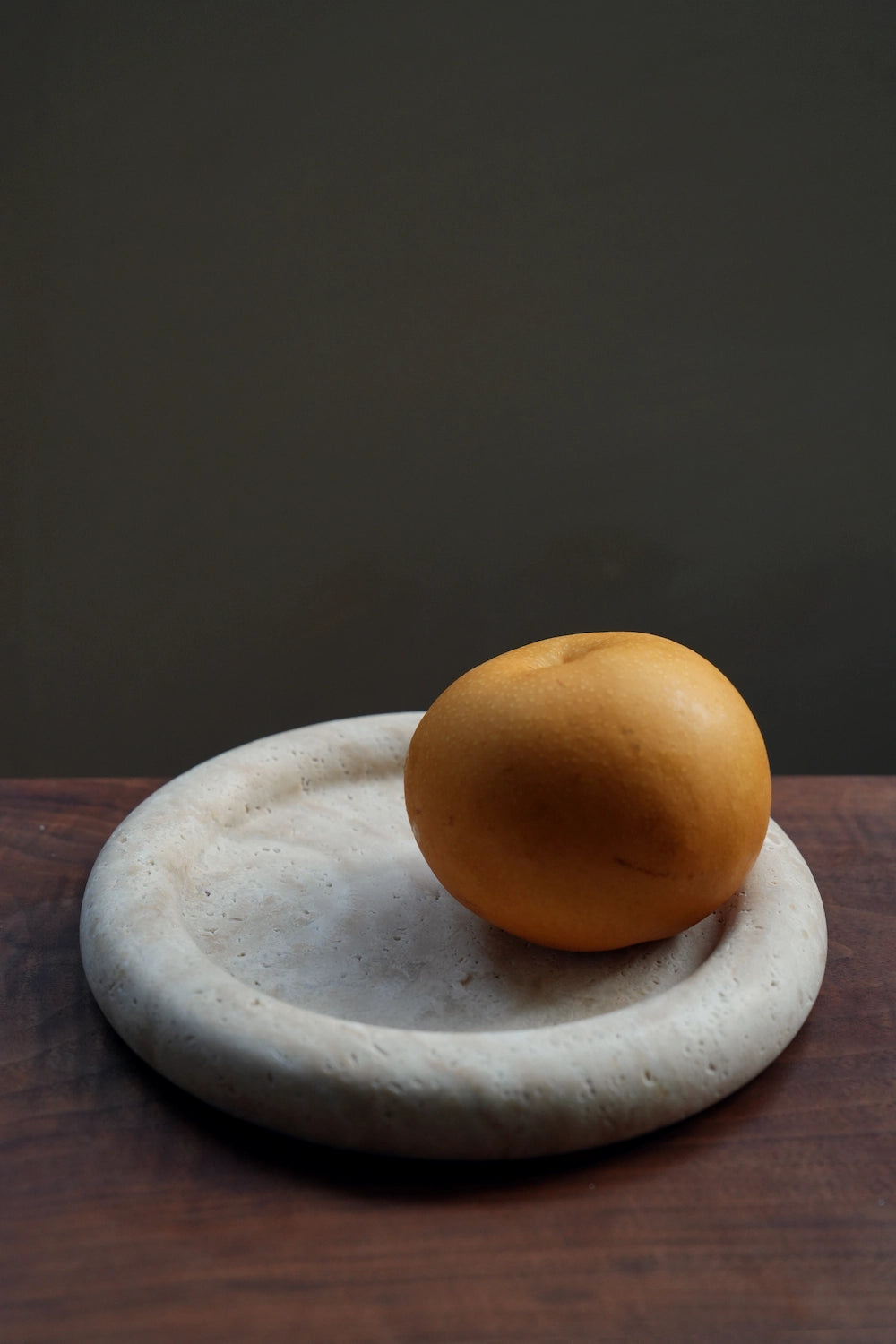 An Asian pear resting in the center of the round travertine dish, highlighting its use as a decorative holder.
