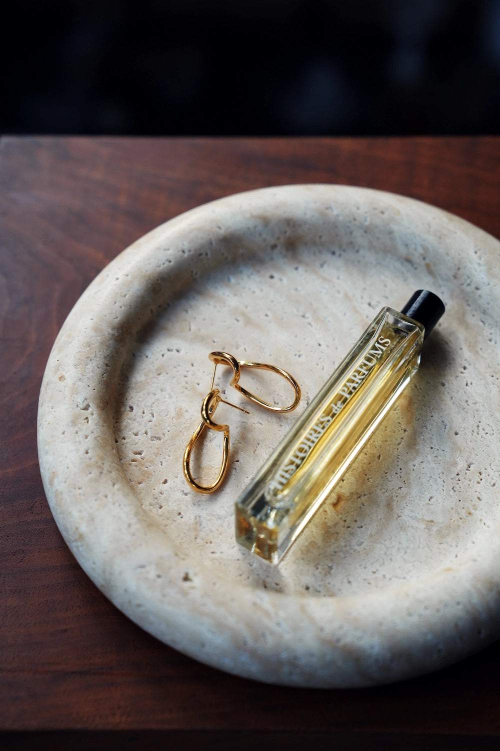 Gold earrings and a bottle of perfume elegantly arranged inside the minimalist travertine jewelry dish.
