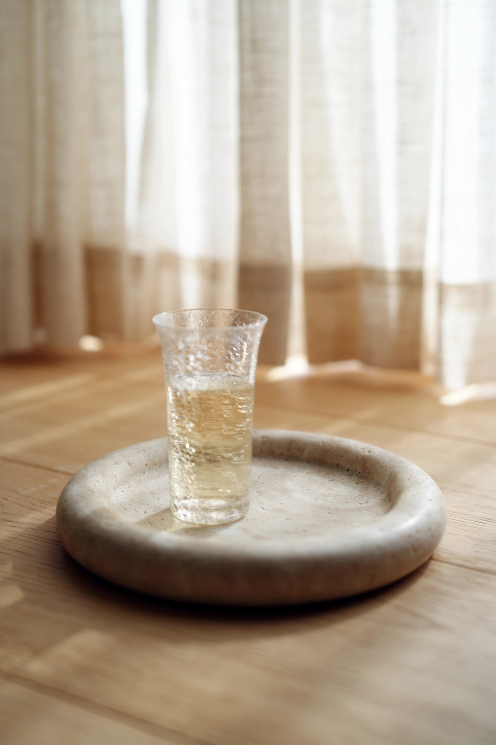 A glass of sparkling water resting on the travertine tray, which serves as a sculptural coaster on a light wood floor.