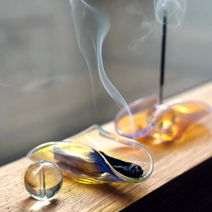 Smoke curling from a Palo Santo stick in a color-shifting glass dish.