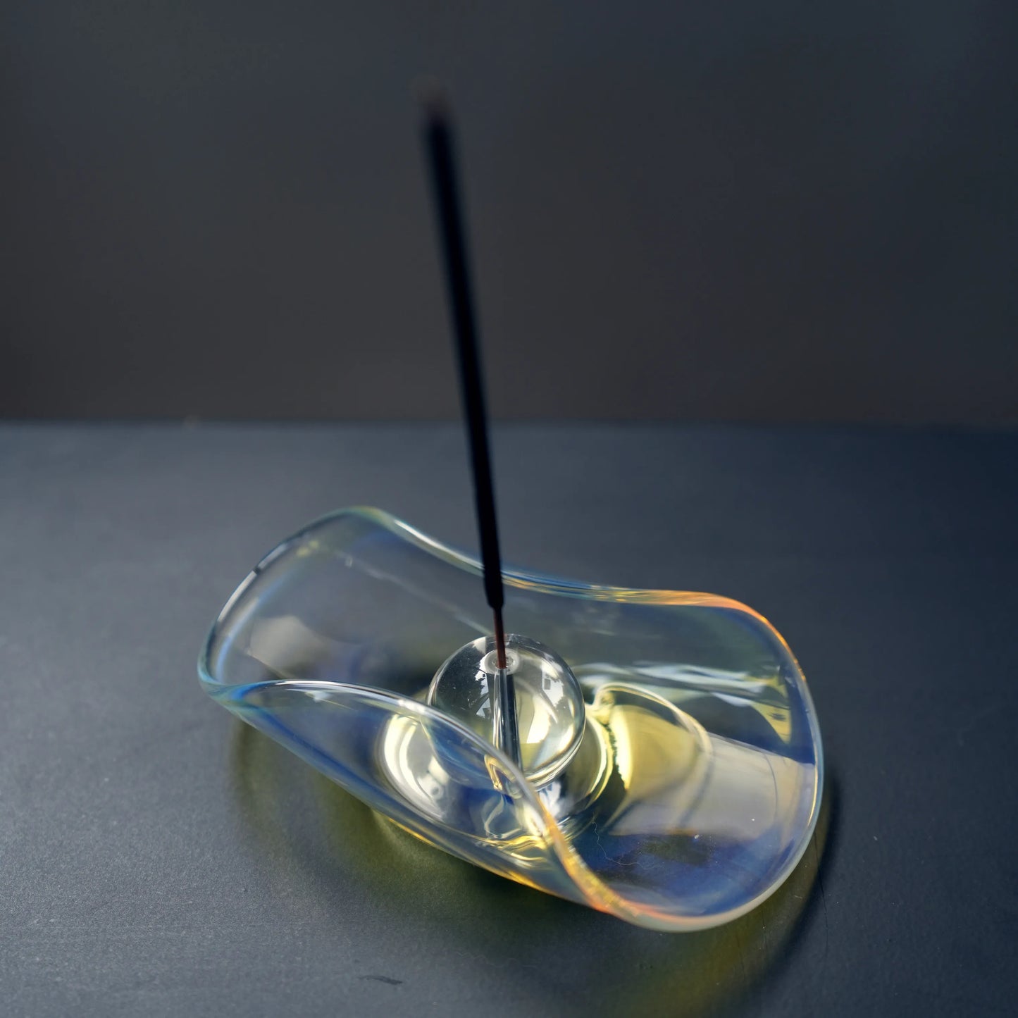 An incense stick burning in the sphere of an iridescent glass holder.