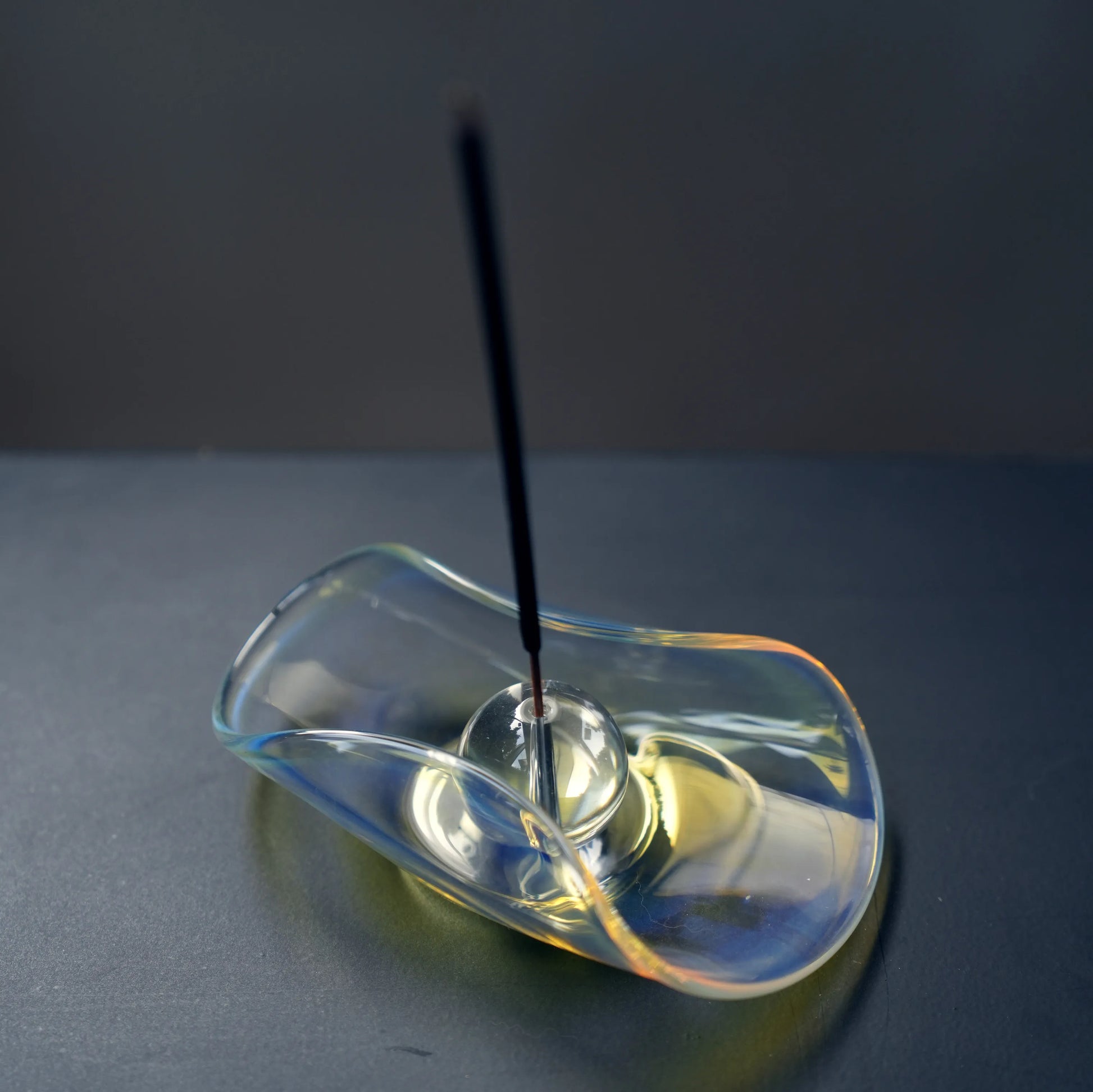 An incense stick burning in the sphere of an iridescent glass holder.