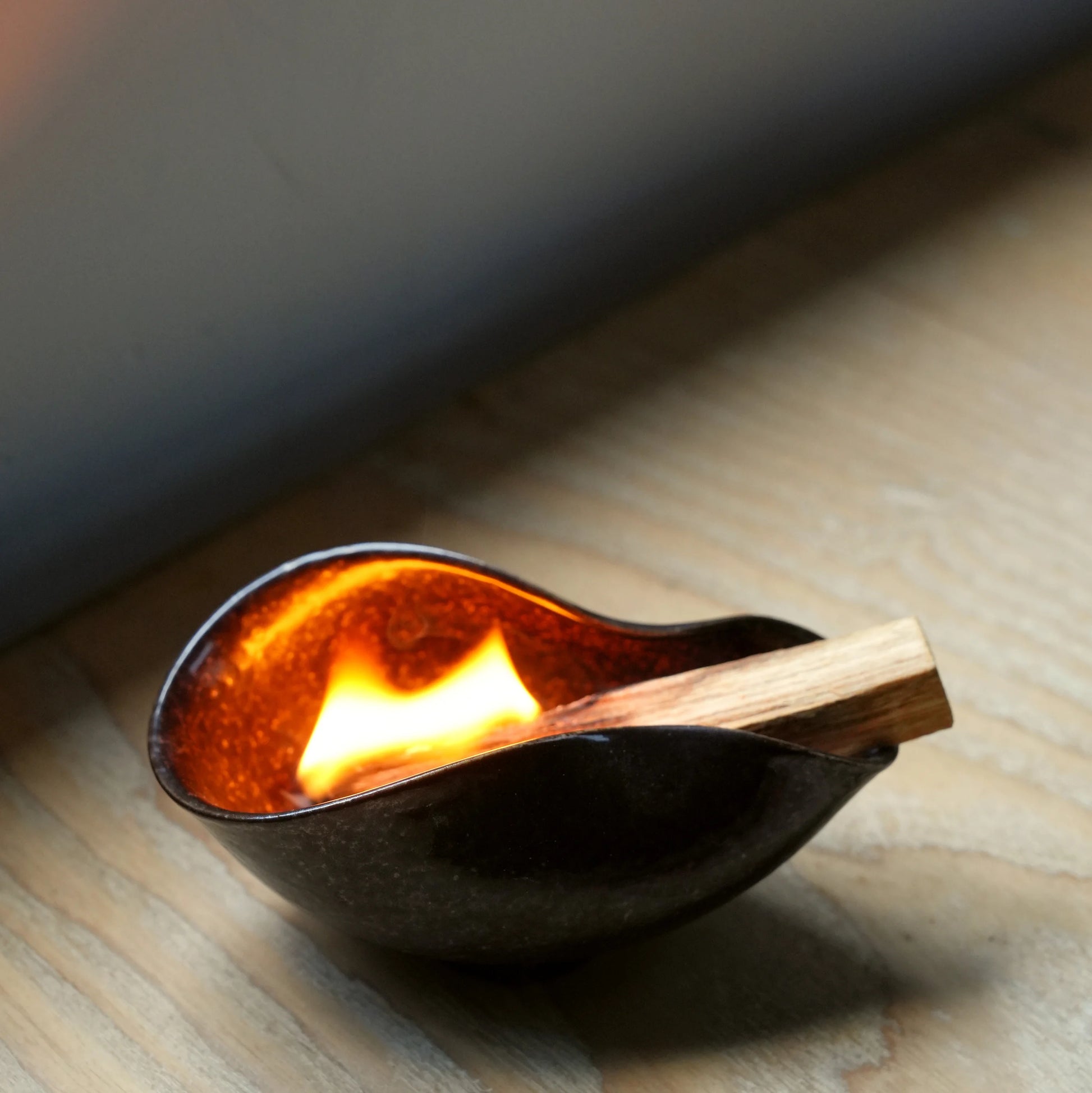 A Palo Santo stick burning brightly inside a dark ceramic ritual bowl.