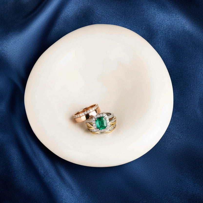 An elegant arrangement of gold and emerald rings resting inside the smooth alabaster bowl, which sits on a rich, navy blue silk cloth.