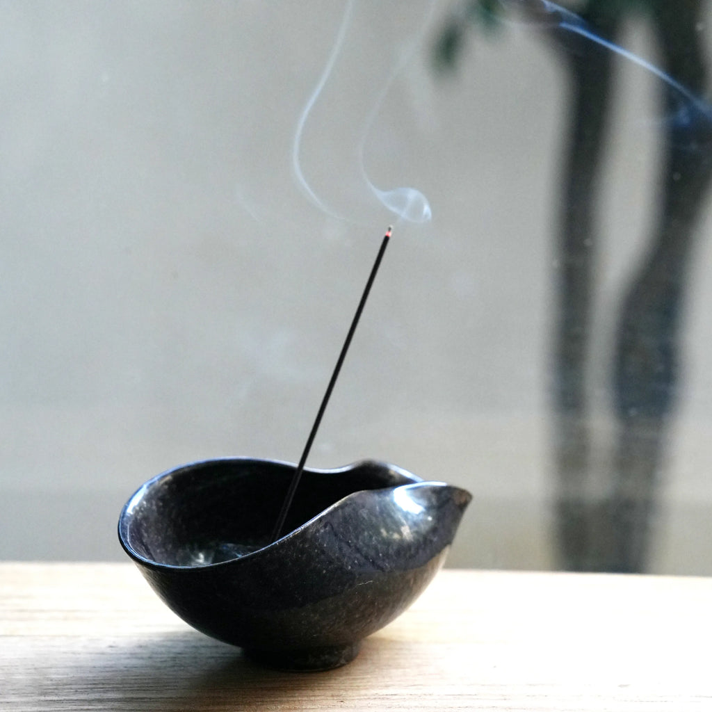 An incense stick burning in a dark ceramic bowl on a wooden surface.
