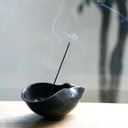 An incense stick burning in a dark ceramic bowl on a wooden surface.