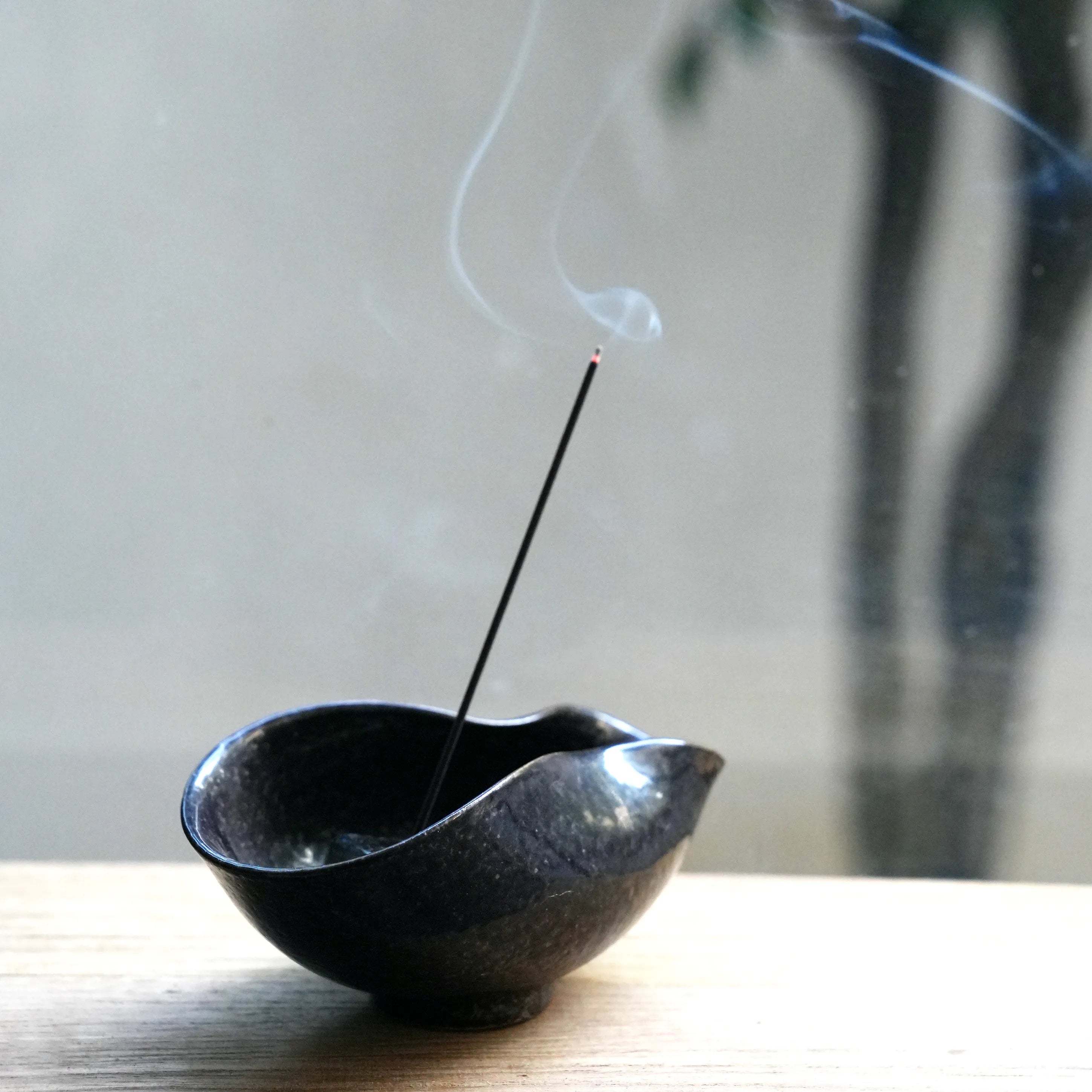 An incense stick burning in a dark ceramic bowl on a wooden surface.