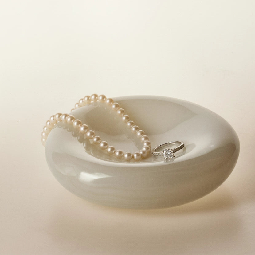 A classic pearl necklace and a diamond ring displayed in the sculptural alabaster catchall dish against a soft cream background.