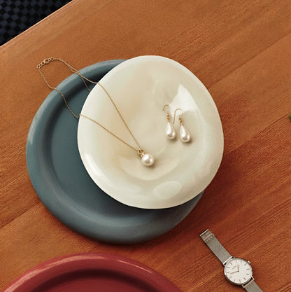 The white alabaster jewelry dish layered on top of other colorful trays, holding a delicate pearl necklace and earrings on a wooden table.