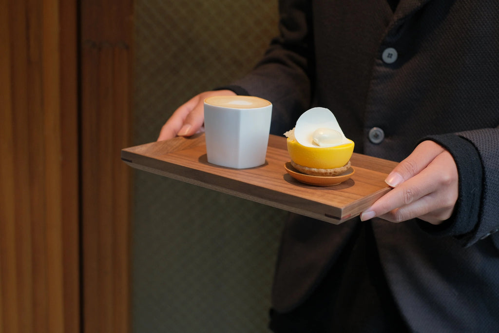 Hands carrying walnut tray with latte and lemon dessert, café