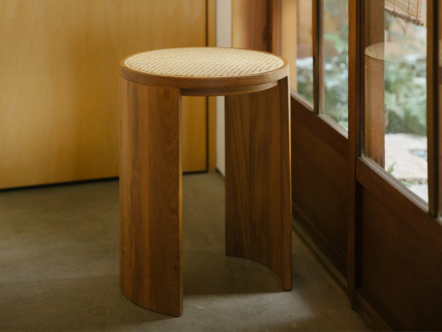 A Japandi-style rattan stool standing on a concrete floor by a doorway.