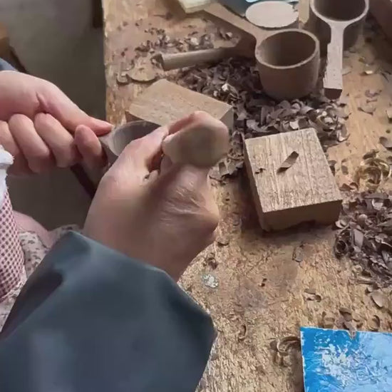An artisan's hands using traditional tools to hand-carve a wooden scoop.