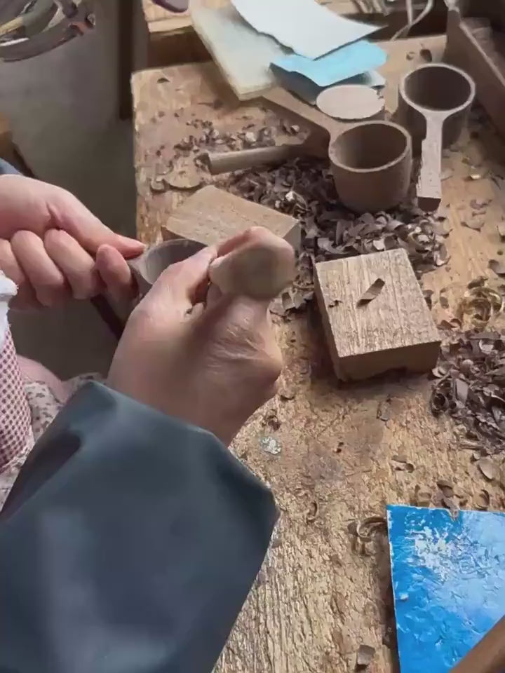 An artisan's hands using traditional tools to hand-carve a wooden scoop.